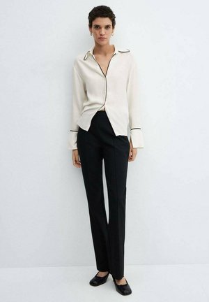 Crème blouse met zwarte bies, lange mouwen en een ontspannen pasvorm, gecombineerd met strakke zwarte broeken. Het model draagt zwarte flats.