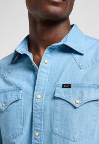 Close-up van een persoon die een lichtblauw spijkershirt draagt met drukknopen en twee borstzakken, waarvan één is voorzien van een klein "Lee" label.