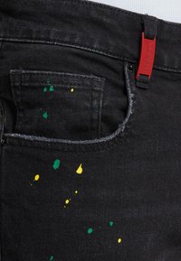 Mustad denim püksid, millel on esi-tasku, kaunistatud kollase ja rohelise värvi tilkadega. Vööl on punane kangast silt.