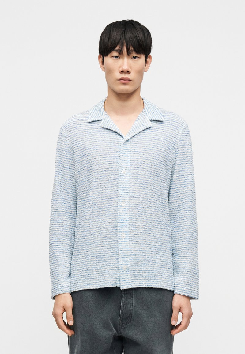 Chemise à rayures bleu clair avec col, en tissu doux. Devant boutonné, manches longues, coupe légèrement ample, portée avec un pantalon gris foncé.