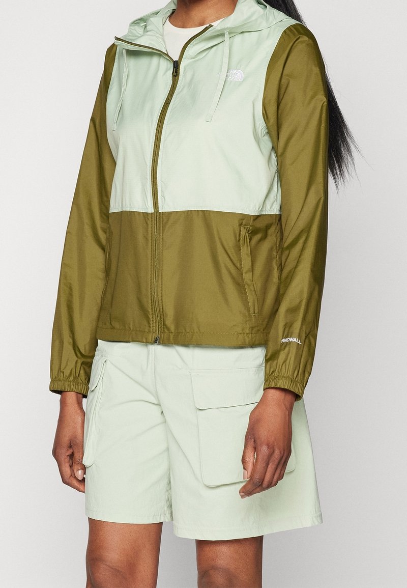 Femme portant une veste à capuche zippée bicolore verte et un short cargo vert clair avec des poches sur un fond uni.