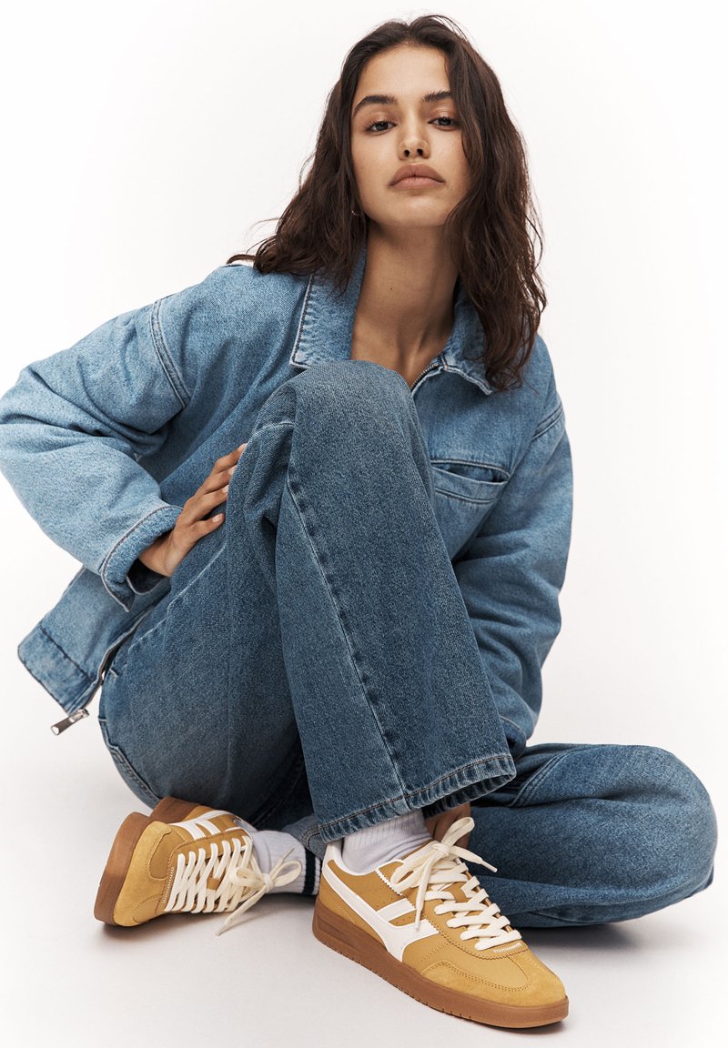 Sneaker mit einem gelb-weißen Design, ausgestattet mit einem strukturierten Material und einer Gummisohle. Kombiniert mit einer blauen Jeansjacke und Jeans.