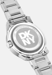 Orologio in acciaio inossidabile argentato con quadrante rotondo e cinturino in metallo spazzolato. Presenta un retro liscio e riflettente inciso con "DKNY". Impermeabile fino a 5 ATM.
