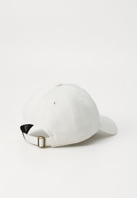 Hvit baseballcaps med buet skjerm, justerbar metallspenne bak, og synlige sømdetaljer på stoffet.