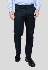 Pantalons de costume noirs, coupe slim, avec une texture lisse. Dotés d'une fermeture à bouton et de poches avant, assortis à des chaussures noires soigneusement cirées.