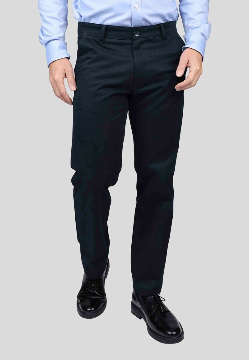 Pantalons de costume noirs, coupe slim, avec une texture lisse. Dotés d'une fermeture à bouton et de poches avant, assortis à des chaussures noires soigneusement cirées.
