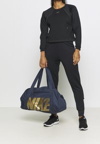 Donna in un completo sportivo nero che tiene una borsa da viaggio blu navy con grande logo dorato "NIKE", in piedi contro uno sfondo bianco semplice.