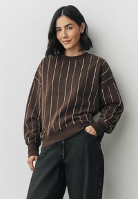 Brązowy sweatshirt z białymi pionowymi paskami, luźny krój, ściągacze na mankietach i dekolcie, połączony z ciemnymi dżinsami. Widoczna miękka tekstura.