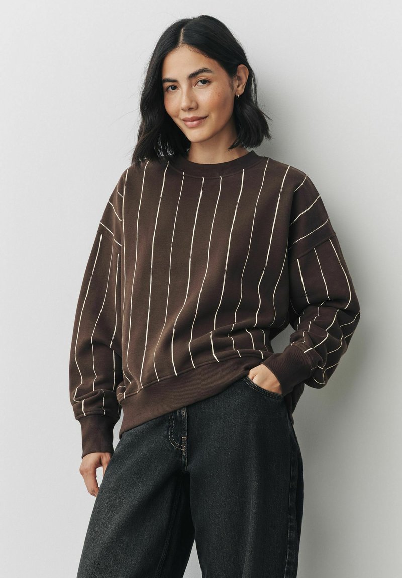 Brązowy sweatshirt z białymi pionowymi paskami, luźny krój, ściągacze na mankietach i dekolcie, połączony z ciemnymi dżinsami. Widoczna miękka tekstura.