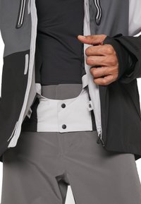 Veste de ski noire et grise avec une poche zippée, une fermeture à pression et des poignets élastiques. Associée à un pantalon léger gris. Tissu texturé.