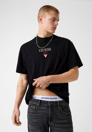 Młody mężczyzna w czarnej koszulce Guess i ciemnych jeansach, lekko podnoszący koszulkę, aby odsłonić białą bieliznę z paskiem Guess Jeans.