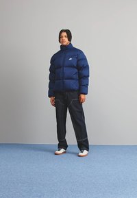 Σακάκι puffer σε ναυτικό χρώμα με ψηλό γιακά και λογότυπο, συνδυασμένο με σκούρες τζιν παντελόνες, λευκά αθλητικά παπούτσια και υφή φόντου.