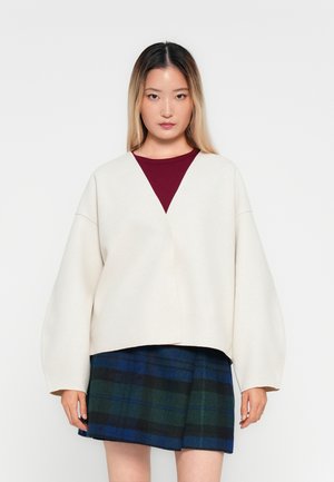 Žena se rovnými ombre vlasy, oblečená v krémovém oversized kabátu přes burgundský top a sukně s modro-zeleným kostkovaným vzorem, stojící před bílým pozadím.
