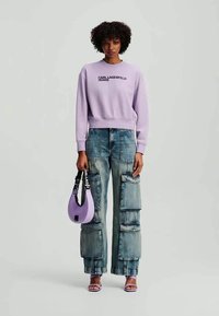 Felpa lilla con logo stampato, jeans cargo oversize con tasche, borsa lavanda e sandali coordinati. Stile streetwear casual.