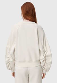 Sudadera color crema con mangas raglán, detalle fruncido en los codos y una cintura de punto. Textura de tela suave.