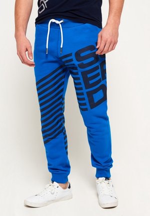 Man draagt blauwe joggers met grote zwarte diagonale strepen en tekst, witte trekkoord en witte sneakers, staand tegen een effen achtergrond.