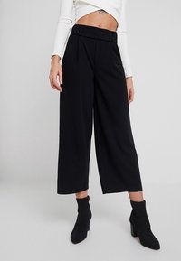 Pantalon noir à jambes larges avec une taille haute et deux boutons. Confectionné dans un tissu lisse. Assorti à des bottines noires à talons montant jusqu'à la cheville.