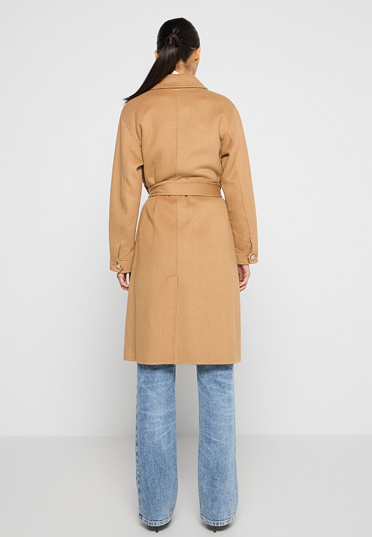 Kobieta z długimi ciemnymi włosami związanymi w kok, ubrana w kamelowy płaszcz z paskiem i jasnoniebieskie spodnie o szerokich nogawkach, stojąca na tle białej ściany.