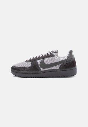Baskets Nike basses avec panneaux noirs texturés, dessus en mesh gris, lacets blancs et semelle noire sur fond gris clair.