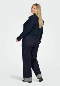 Kvinna som ler över axeln, iförd en mörkblå denimjacka och matchande vida jeans med uppvikta fållar samt svarta skor.