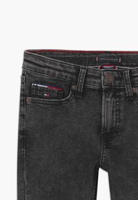Los vaqueros de denim negro cuentan con un diseño clásico de cinco bolsillos, herrajes de cobre, costuras rojas y azules, y una etiqueta de marca en la cintura.