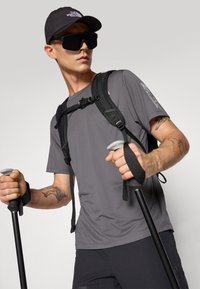 T-shirt grigio traspirante, zaino nero, cappellino nero, occhiali da sole neri oversize, in mano pali da trekking con impugnature grigie e cinturini neri.