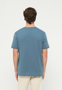 Görögbarna göndör hajú férfi, egyszerű kék rövid ujjú pólót és bézs nadrágot visel, háttal áll, egyszínű háttér előtt.