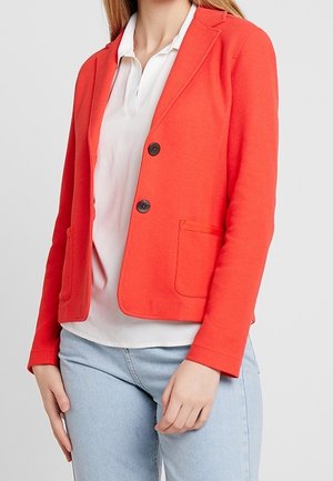 Vrouw die een feloranje blazer met twee knopen draagt over een wit overhemd met kraag en lichtblauwe spijkerbroek, staand tegen een effen achtergrond.