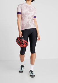 Ljust rosa cykeltröja med vita och lila detaljer, svarta cykelbyxor, hållandes en röd och svart hjälm, iförd sportskor och vita strumpor.