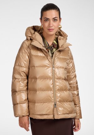 Vrouw met donker haar die gouden creolen draagt en een glanzende bruine donsjas over een gebloesd hemd op een witte achtergrond.