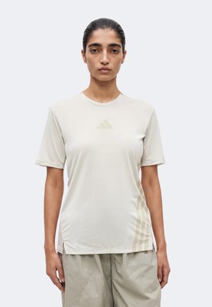 Žena s uhlazenými vlasy, která má na sobě světle béžové krátké tričko Adidas a volné béžové kalhoty, stojící před jednobarevným pozadím.