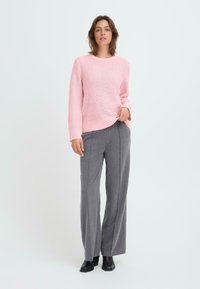 Heller rosa gestrickter Pullover mit einem strukturierten Muster, kombiniert mit grauen weiten Hosen und schwarzen Stiefeletten. Schlichtes, modernes Design.