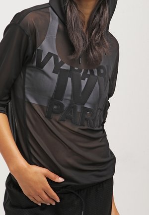 Osoba ubrana w czarną przezroczystą bluzę z kapturem z napisem „IVY PARK” oraz czarne sportowe spodenki, ręka spoczywa na biodrze.