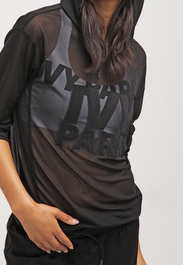 Personne portant un sweat à capuche noir transparent avec le texte "IVY PARK" et un short de sport noir, main reposant sur la hanche.