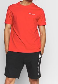 Rotes Baumwoll-T-Shirt mit kurzen Ärmeln und Rundhalsausschnitt, mit einem weißen "Champion"-Logo auf der Brust. Schwarze Shorts mit Logo-Akzent.