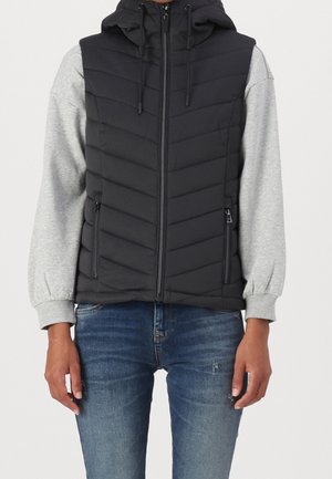 Schwarze gesteppte ärmellose Kapuzenweste mit Reißverschluss und Reißverschlusstaschen, getragen über einem hellgrauen langärmligen Sweatshirt und blauen Jeans.