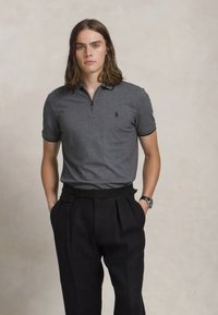 Γκρι πόλο μπλούζα με κοντά μανίκια και κολάρο με φερμουάρ, συνδυασμένη με μαύρο πλισέ παντελόνι. Η πόλο διαθέτει ένα μικρό λογότυπο στο στήθος.