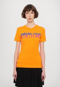 Oranžové bavlněné tričko s nápisem "VERSACE JEANS COUTURE" v purpurové a modré barvě přes hruď; krátké rukávy, přiléhavý střih.