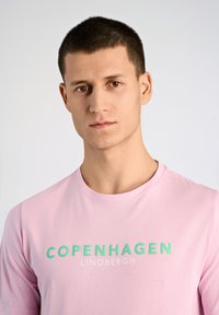 Vaaleanpunainen t-paita, jossa on mintunvihreällä teksti "COPENHAGEN" ja valkoisella "LINDBERGH". Pyöreä pääntie, sileä rakenne, lyhyet hihat.