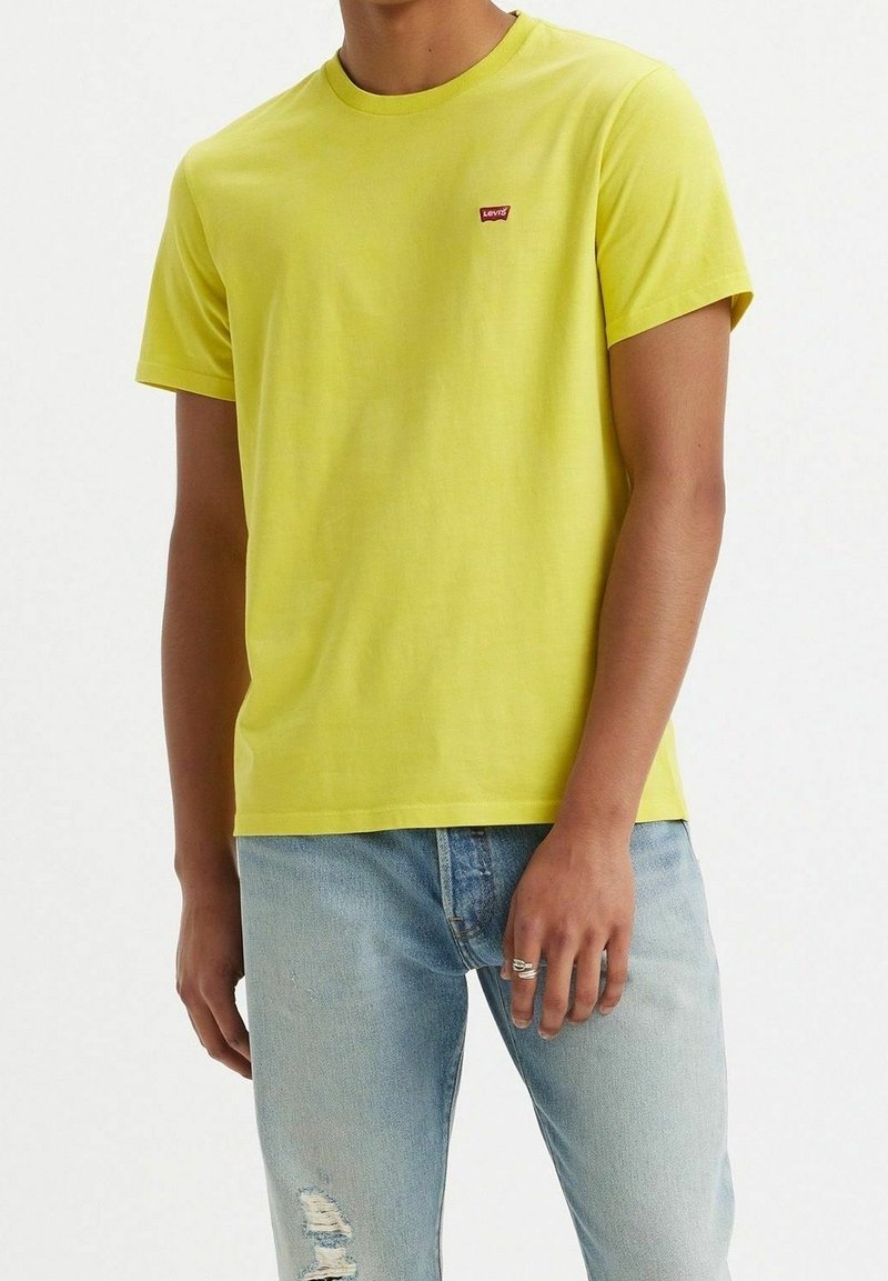 Homme portant un t-shirt Levi's jaune vif et un jean bleu clair déchiré, avec une bague en argent à la main droite.