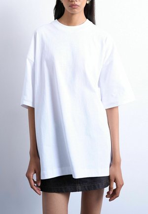 Woman wearing an oversized plain white T-shirt with elbow-length sleeves and a black skirt against a white background.