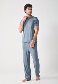 Man wearing a blue-gray short-sleeve shirt and matching pants, standing confidently with one hand on his neck against a plain backdrop.
