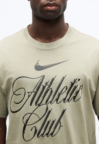 Homme portant un t-shirt Nike vert clair avec l'inscription noire « Athletic Club » et le logo Nike Swoosh à l'avant.