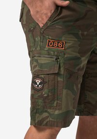 Shorts cargo camouflés dans des tons de vert avec un patch brodé, un détail '086' orange et plusieurs poches, y compris une poche zippée. Tissu texturé.