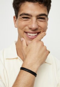 Pulsera de cuero negro llevada en una muñeca, con dos bandas entrelazadas. La persona lleva una camisa de color claro y un anillo dorado.