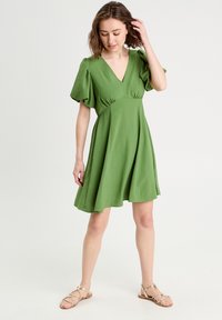 Robe verte à manches courtes avec un décolleté en V profond, corsage ajusté et jupe évasée. Tissu doux et texturé avec des manches bouffantes structurées.