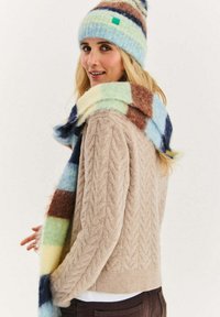 Beżowy sweter z grubym splotem, miękka niebiesko-brązowa czapka w paski oraz wielokolorowy szalik w kratę w delikatnych pastelowych odcieniach.