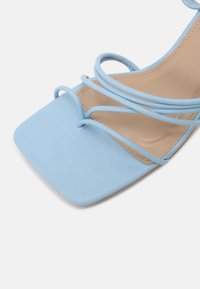 Sandale à talon bleu clair avec un bout carré, dotée de fines lanières incurvées et d'une texture lisse. Semelle intérieure beige douce visible.