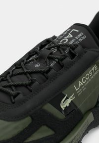Primo piano di una sneaker Lacoste nera e verde con lacci a fantasia e testo bianco del marchio sul lato e sulla linguetta.