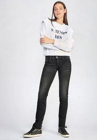 Jean slim foncé associé à un sweat-shirt gris clair avec un texte bleu marine. Le modèle porte des baskets noires scintillantes. Tissu lisse, coupe ajustée.